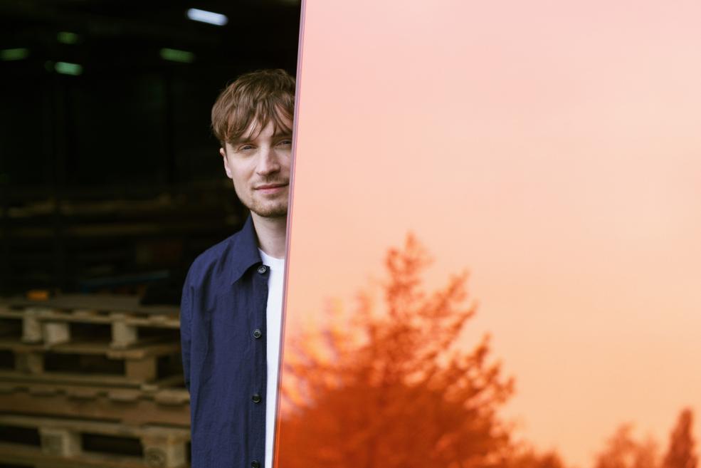 Germans Ermičs stāv pie orandžas stikla sienas, kurā atspīd koka siluets. 