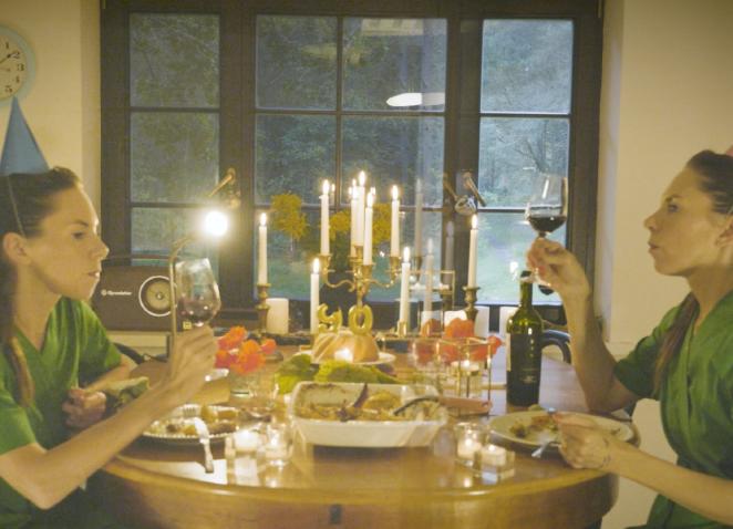 Fotogrāfija ar Katrīnu Neiburgu, kura viena sēž pie galda ar savu atspulgu