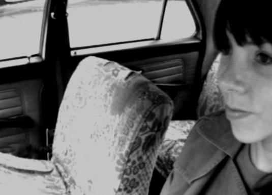 Katrīna Neiburga sēž mašīnā pie autovadītāja stūres. 